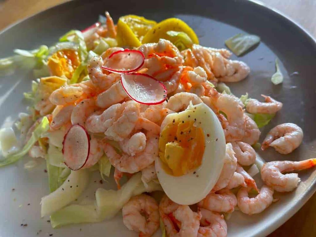 A close up photo of a shrimp sandwich overflowing with pink shrimps, green salad, eggs and raddish