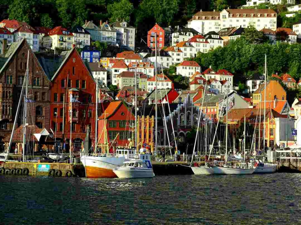 A port area of a town with boats on the water, charming colorful old buildings surrounding the port, and white old houses clinging to the hillside stretching out of town on a sunny summer day with lots of trees and greenery