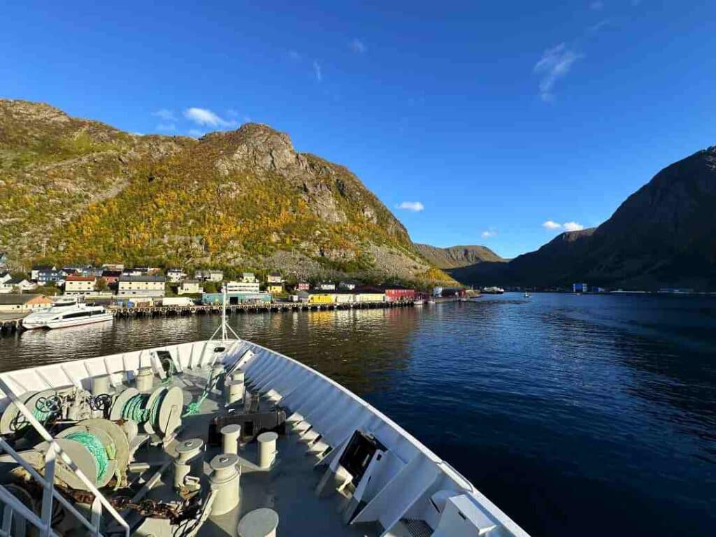 The white bau of a big ship approaching the port in a small Arctic fishing village on dark waters on a bright sunny summer day with blue skies, and green mountains and hills behind the village