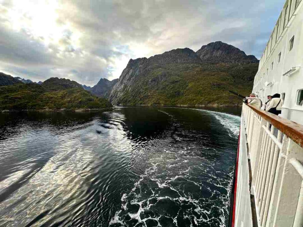 A ship has just left a narrow fjord behind, and you see the ship side and the aft white water train into the fjord canyon between steep mountains