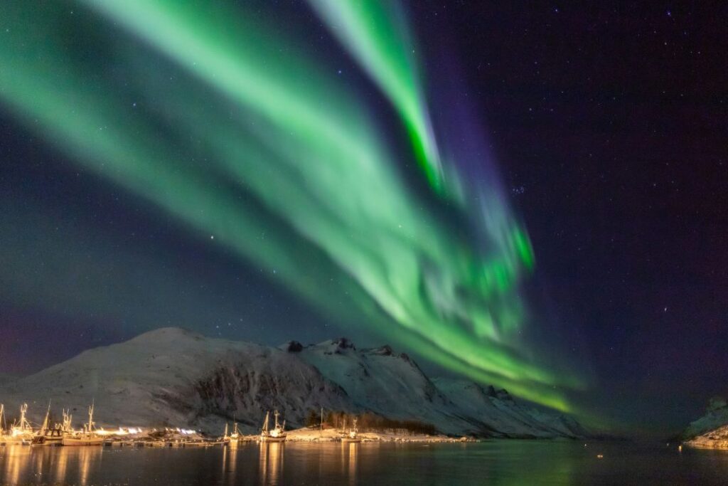 A huge green wave of Aurora Borealis, the northern lights, across a dark blue sky over a snow covered mountain and a small fishing village 