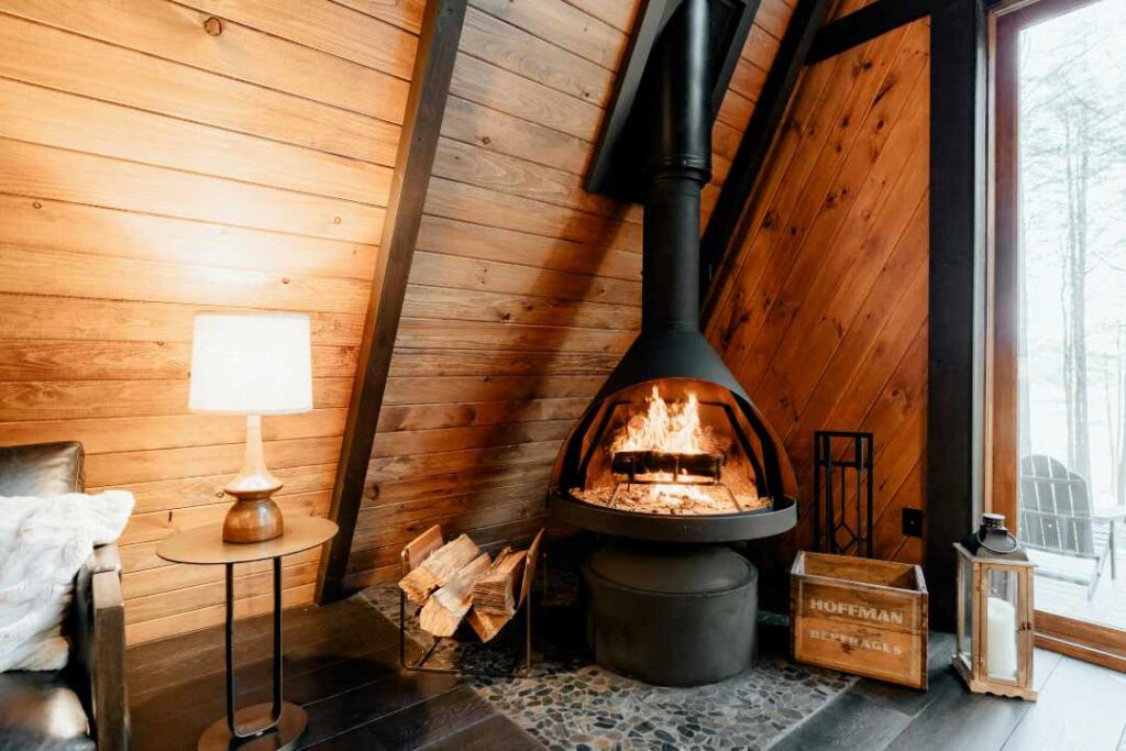 A modern fireplaces crackling with fire in a wooden cabin with warm lighting, and cool daylight from the winter outside coming through the tall wide window bringing nature into the home