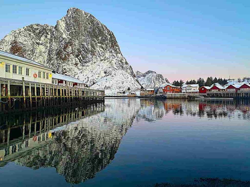 A quiet harbor on a calm winter day in Lofoten, surrounded by charming traditioal wooden cabins, in front of a steep majestic snow covered mountain reflecting in the calm water. 