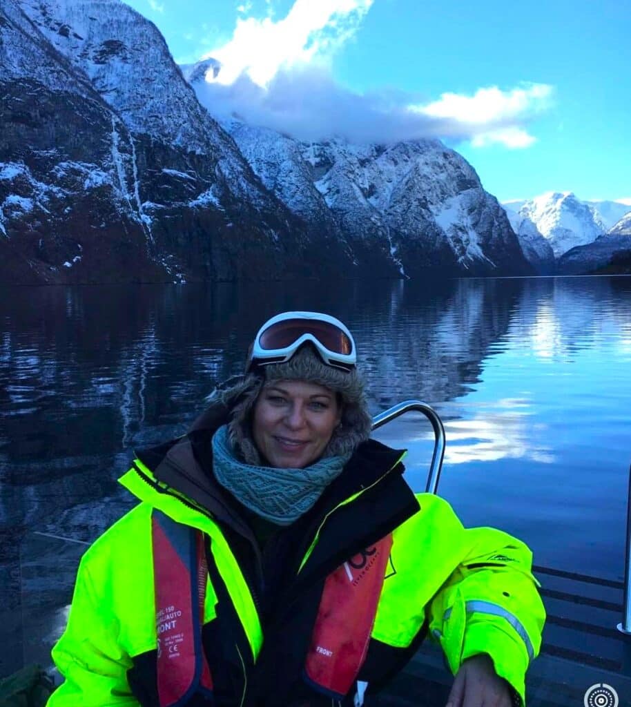 Woman smiling wearing a thick survival suit, hat, and goggles, standing on a boat on the fjord below majestic steep mountains in western Norway, ready for a RIB safari