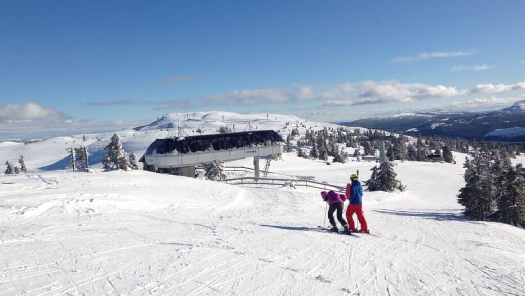 Two people standing on the top of the slopes in a ski resort, with bright white dry snow in the slopes under a bright blue sky on a clear beautiful winter day