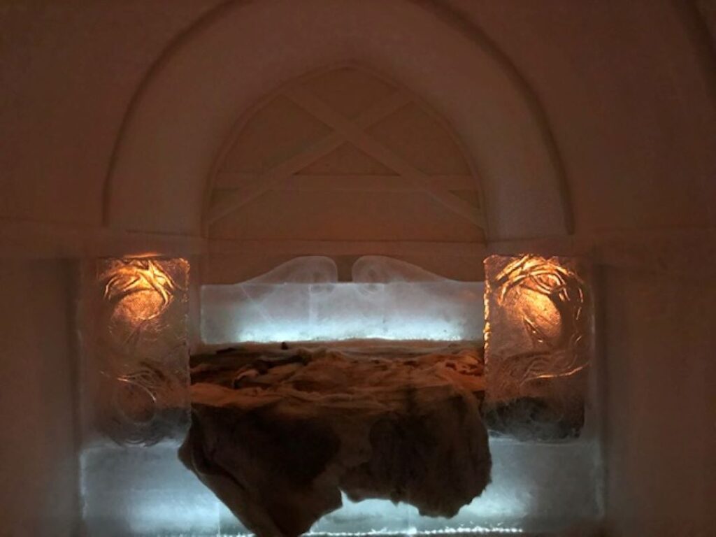 The interior of an ice hotel in Alta, Norway, with a bed made of ice, covered in reindeer fur, with elaborate ice sculptures around it and a warm light