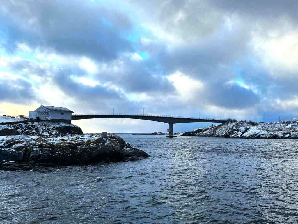 One of the impressive long bridges that connects the many islands in the Lofoten archipelago in northern Norway, crossing the dark sea while snow is on the ground on either side under a clouded sky. 