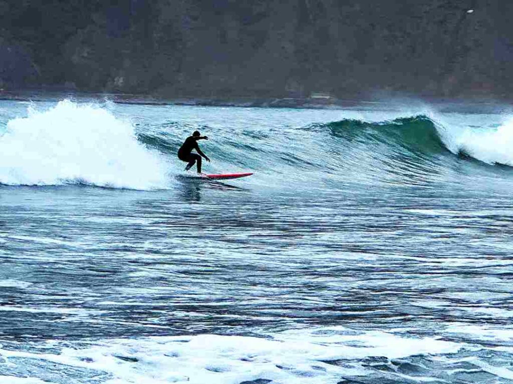 A surfer in a dark suit riding a wave in the dark green Arctic water of the Lofoten Islands in winter. 