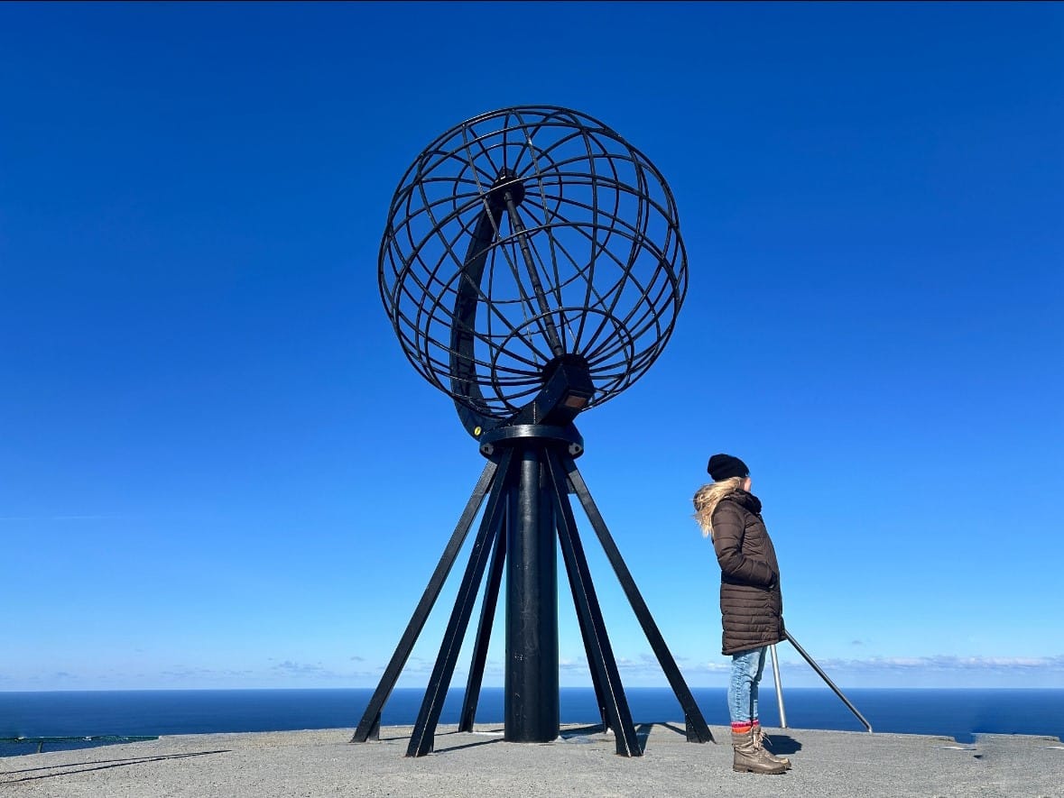 Woman standing beside a globe shaped monument at the North Cape plateau in Norway, under a deep blue sky, in front of the vast ocean staring into the distance - where there is nothing but sea.