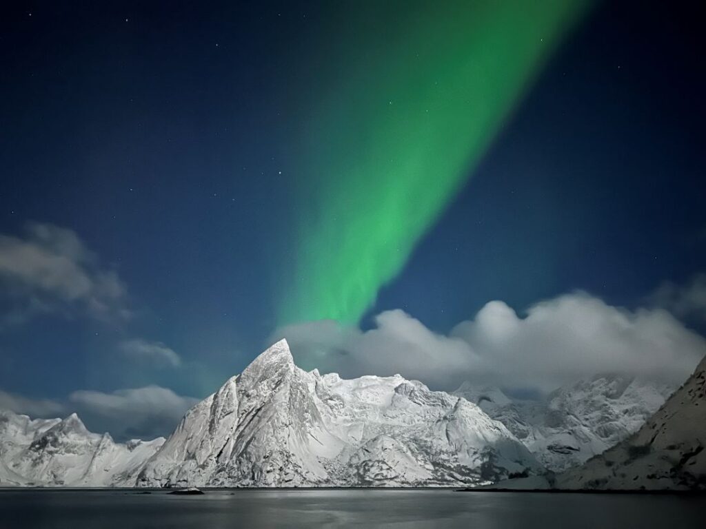 Northern Lights over Lofoten Norway, with green light waves over the dark blue sky over snow covered majestic mountains across the vast fjords