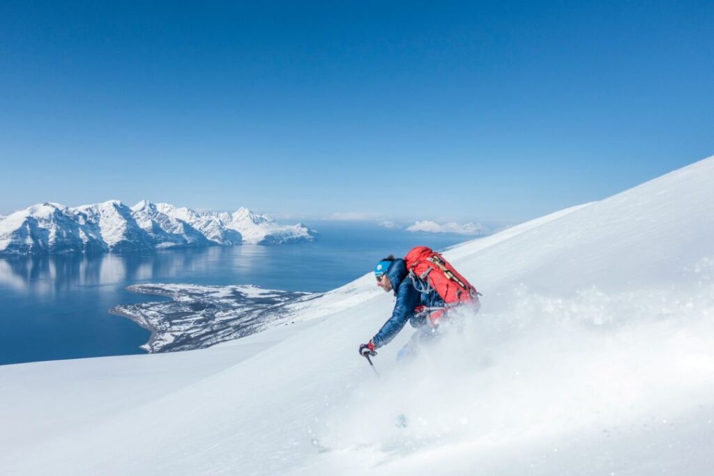 Backcountry skier heading down a mountain with white powder snow on a sunny winter day under a deep blue sky, with the vast fjords and mountains of Norway below him. 