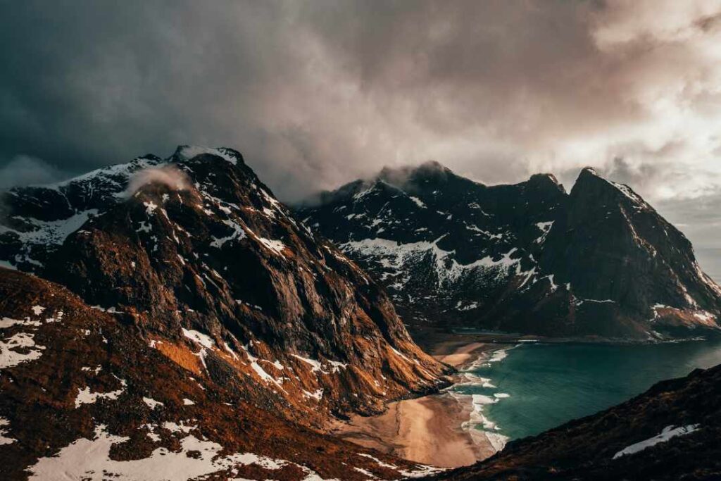 Impressive dark mountains with patches of snow glowing from the sunlight from the sun outside of the photo, hovering over a tiny golden sandy beach at the foot of the mountain by the dark green water. 