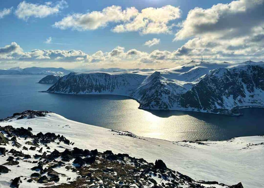 The sun is back in Northern Norway in February, and the snow reflects the bright sun from a pale blue sky, from the top of a mountain with iconic and infinite views of the vast fjords of Northern Norway