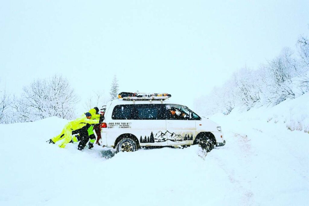 People in bright yellow clothes pushing a car that is stuck in the snow on a small road in the middle of a forest