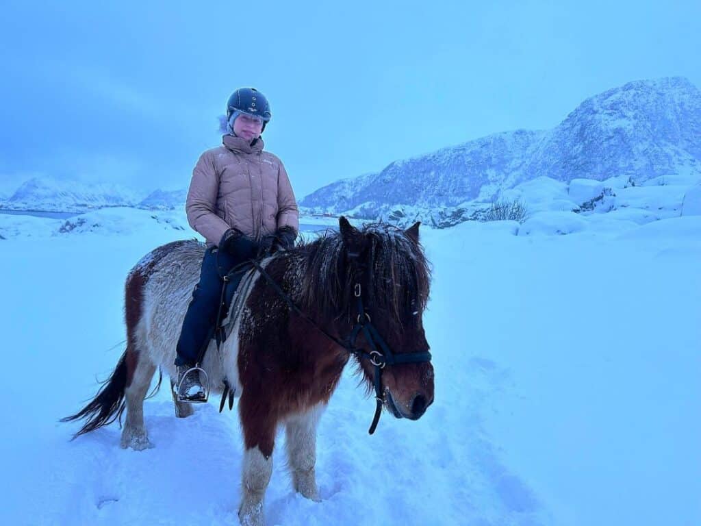 A woman on a horse in the middle of a snow storm in the blue winter light in Lofoten Norway, under a white cloudy sky with vast mountains in the distance