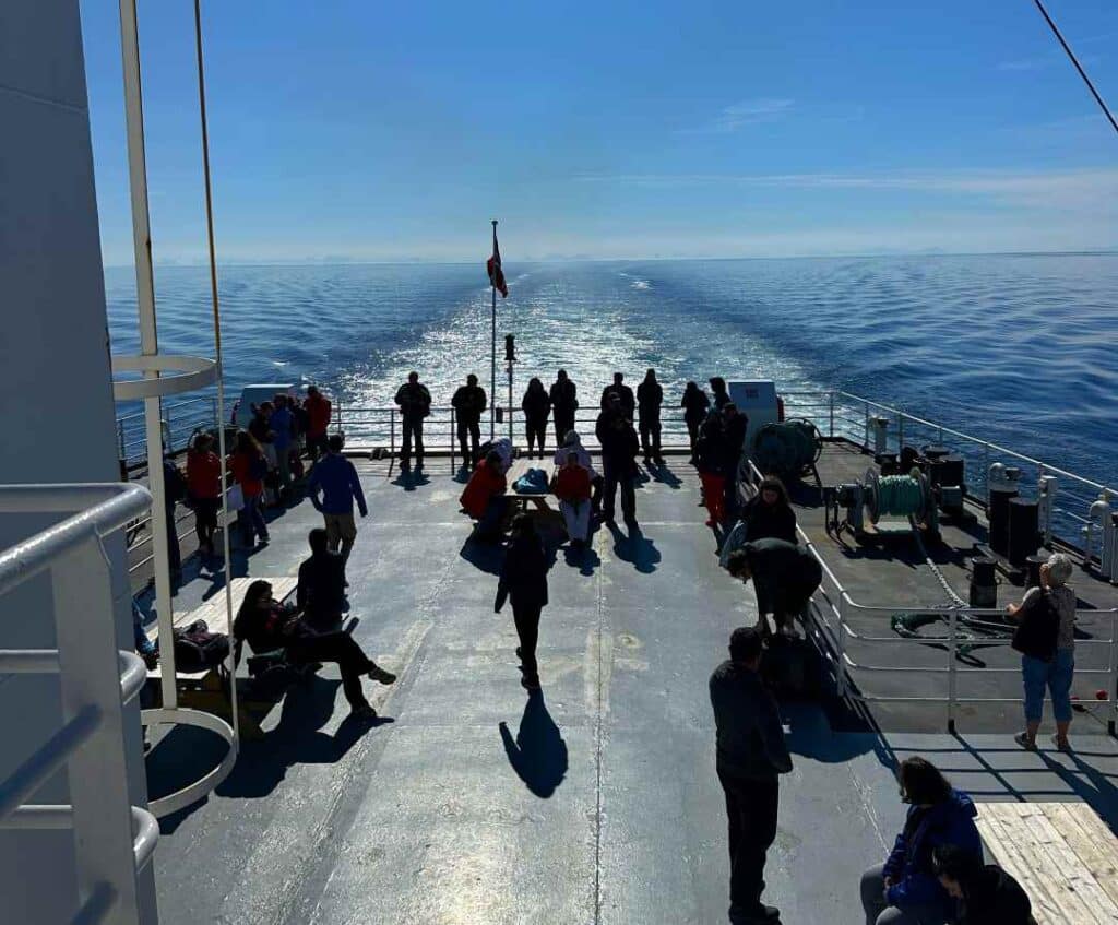 People standing in the sun on the aft deck of a ferry, looking towards the horizon over deep blue waters under a clear blue sky on a bright summer day.