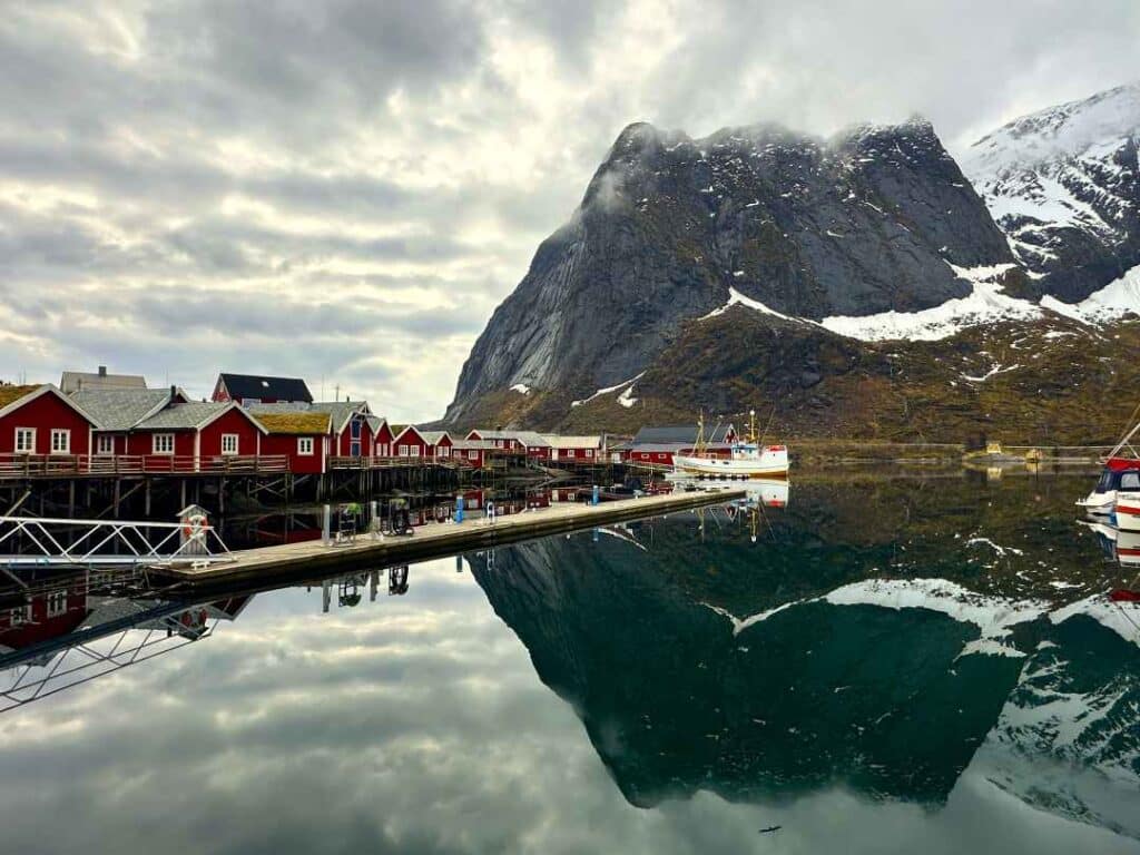 Red fishermans cabins next to a blank harbor on a cloudy day with sunlight piercing through the layer, with majestic mountains with snow covred peaks in the background mirrored in the bland sea