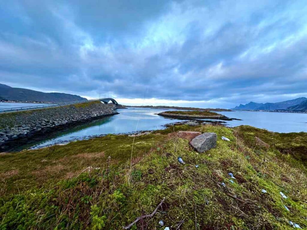 A beautiful curved bridge over a blank fjord under bluish clouds and mountains in Lofoten Norway, with lush greenery in the forefront
