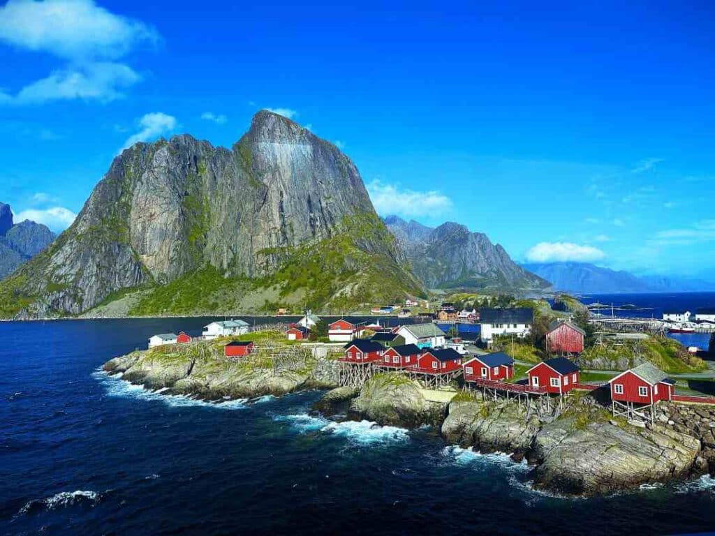One of the most photographed places in the world is this iconic mountain in Reine, Lofoten. Towering over the dark sea behind a small island scattered with red fishermans cabins, under a deep blue sky with bright green bushes, and the Lofoten mountains stretching out in the distance.