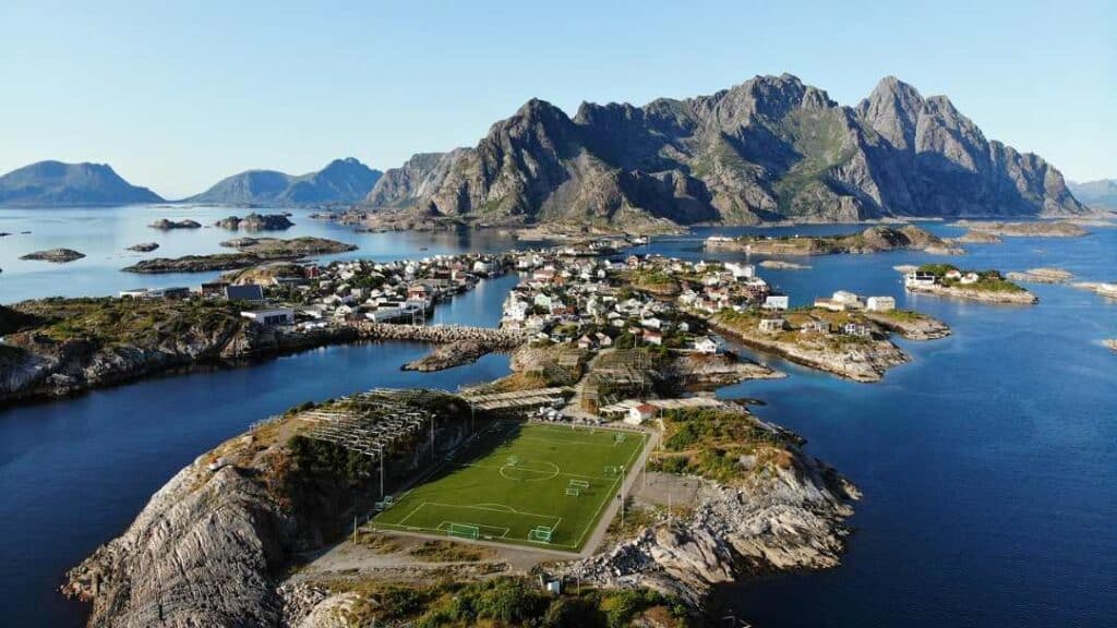 The famous green football field in Henningsvær Lofoten, covering almos an entire little island surrounded by deep blue arctic waters. The fishermans village in the distance, surrounded by the majestic Lofoten mountains under a pale blue sky.