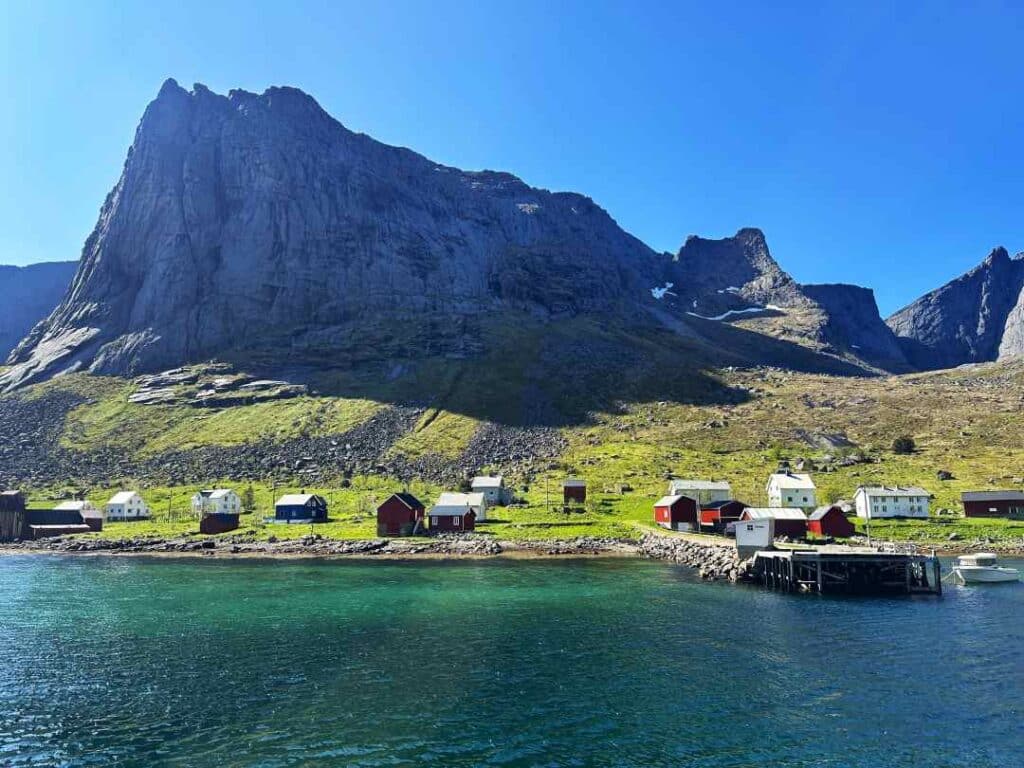 The ferry dock in Kirkefjorden with emerals waters in Lofoten, on a bright sunny day, with scattered red and white houses on the green hills below a majestic grey mountain under the deep blue sky