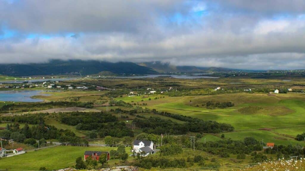 Green fields and small forests with scattered white houses and green hills in the distance under a cloud dotted sky with streaks of blue here and there outside of Leknes in Lofoten Norway