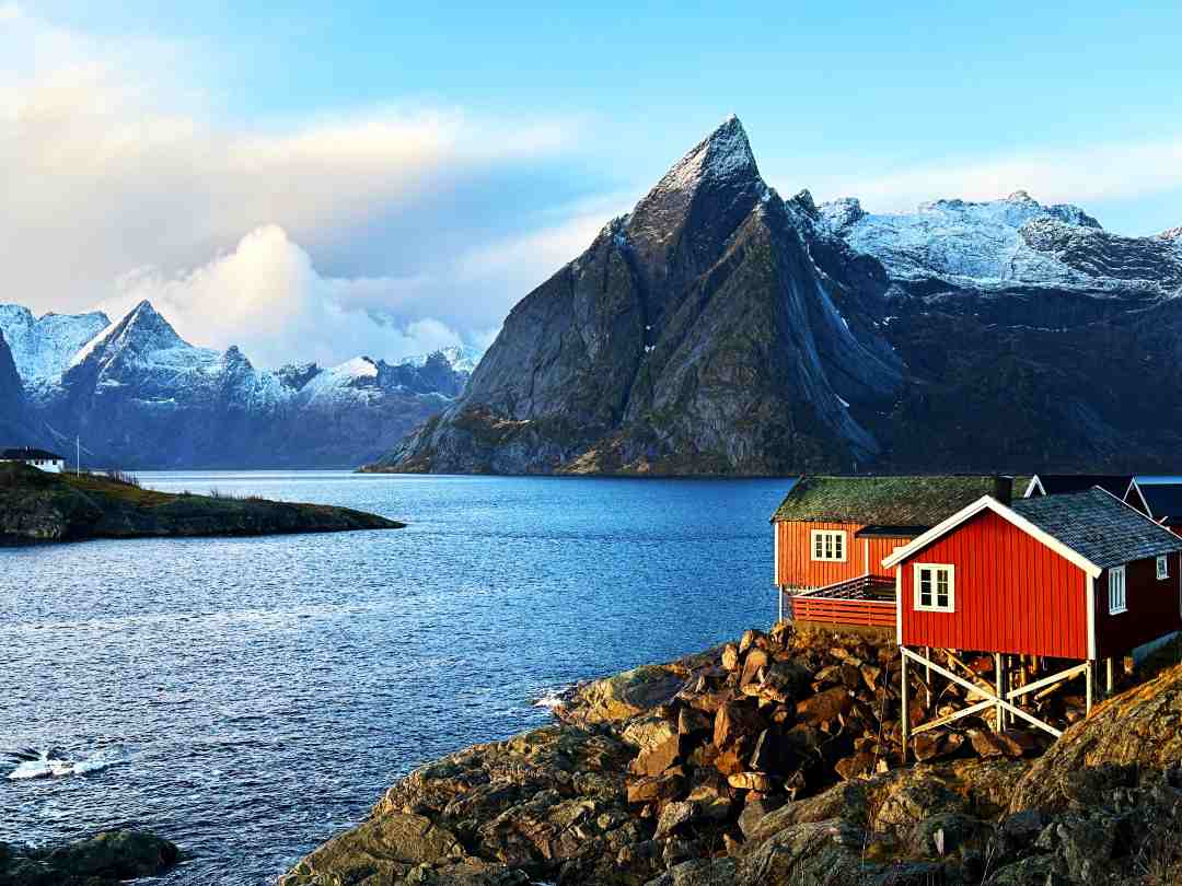 Traditional red fishermans cabins nestled by the fjords under majestic towering dark mountains under a clear blue sky