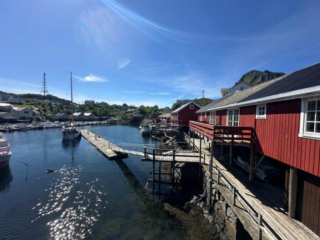 A quiet harbor surrounded by red fishermans cabins and wooden docks with a few boats tied to the pier. Blue skies above, and everything bathed in sunlight, on a bright summer dat, with a green hill in the distance across the water.