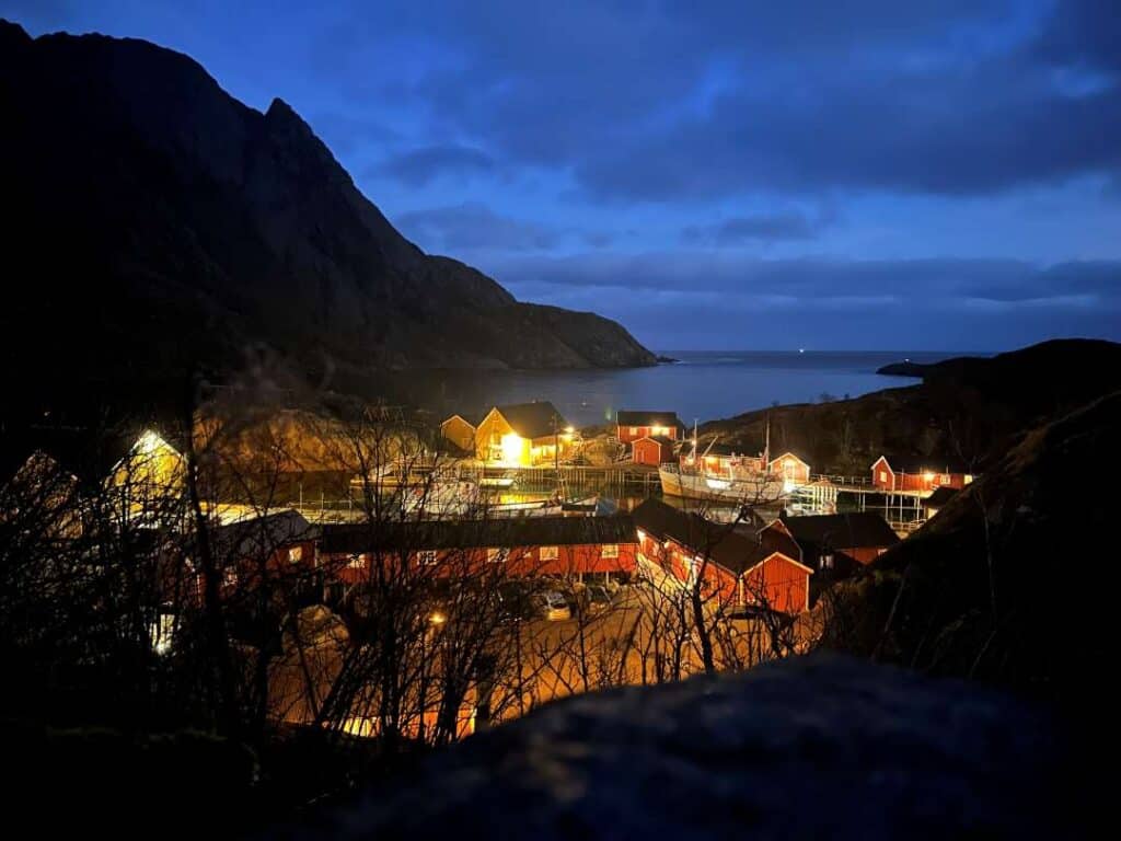 Red fishermans cabins in Nusfjord village in Lofoten during the Polar Night, with some bluish light lingering in the sky over dark mountains and golden lights in the small village