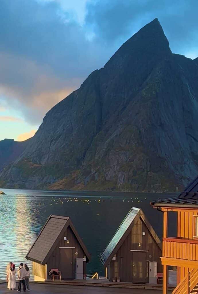 Two wooden saunas by the fjord in front of a massive grey mountain, the sun setting behind the mountain, seafulls on the water and a golden light over it all