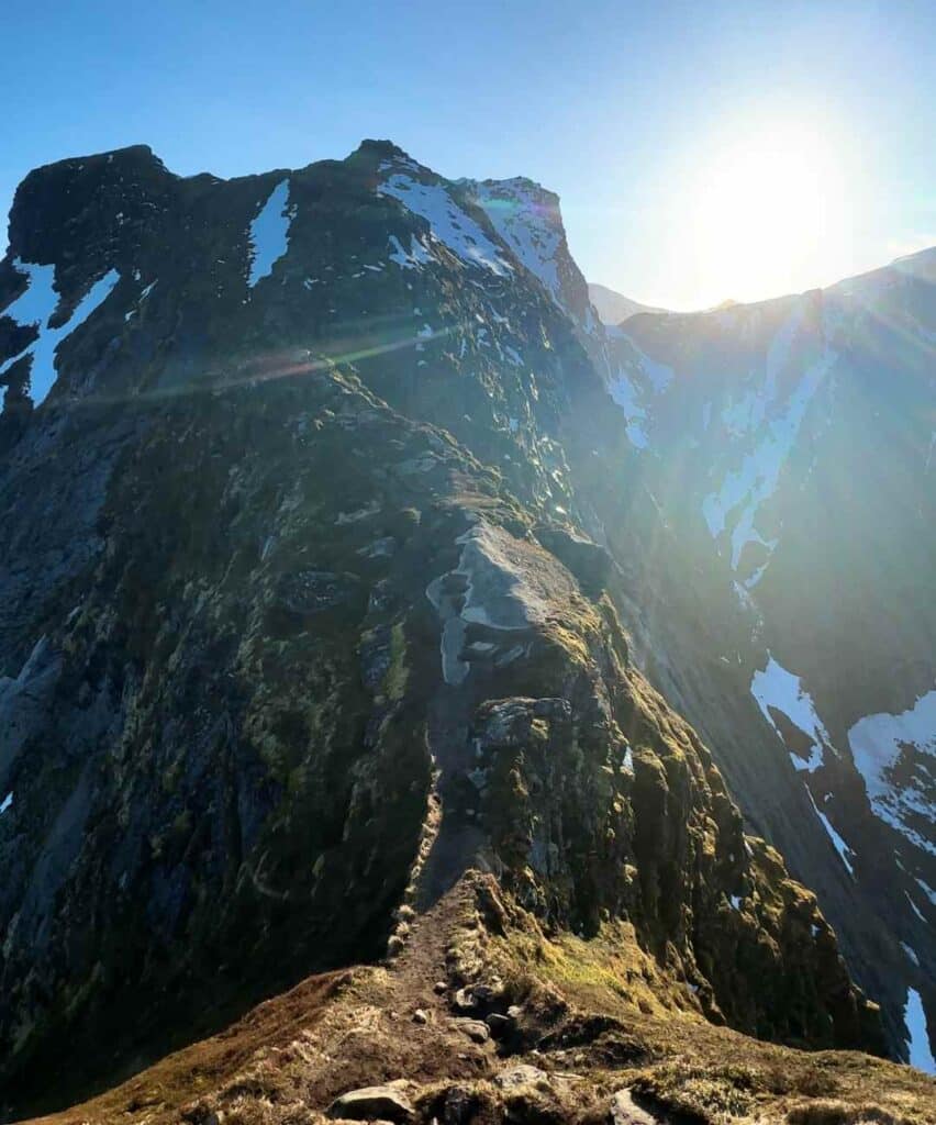 The narrow mountain ridge on Reinebringen mountain in Lofoten, on the way to the actual summit, with snow patches here and there, and the glowing sun in distance leaving a glowing light along the ridge