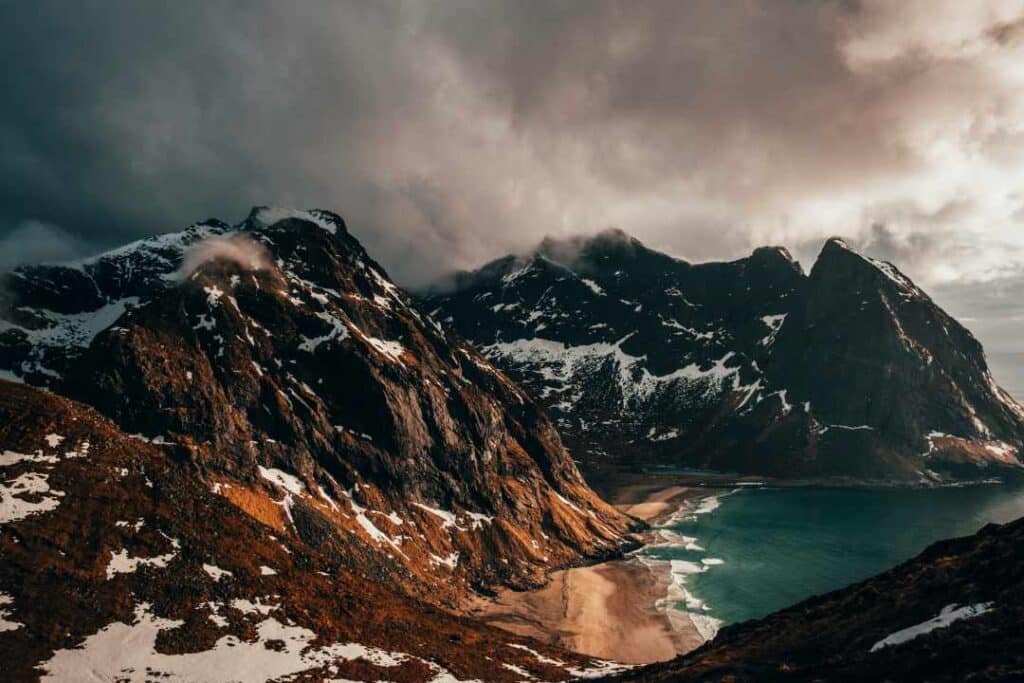Spectacular views of show patched mountains and the golden sandy Kvalvika beach next to dark bluish green waters from Ryten Mountain in Lofoten Norway, under a dramatic greyish clouded sky.