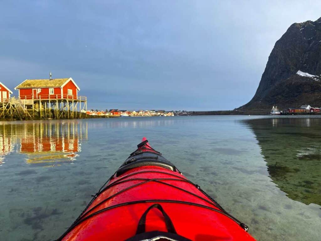 A red kayak gliding over shallow crystal clear waters over a sandy bottom in Lofoten, with red fishermans cabins glowing in the summer sunlight in the distance under a blue sky, with a majestic dark mountain in the background