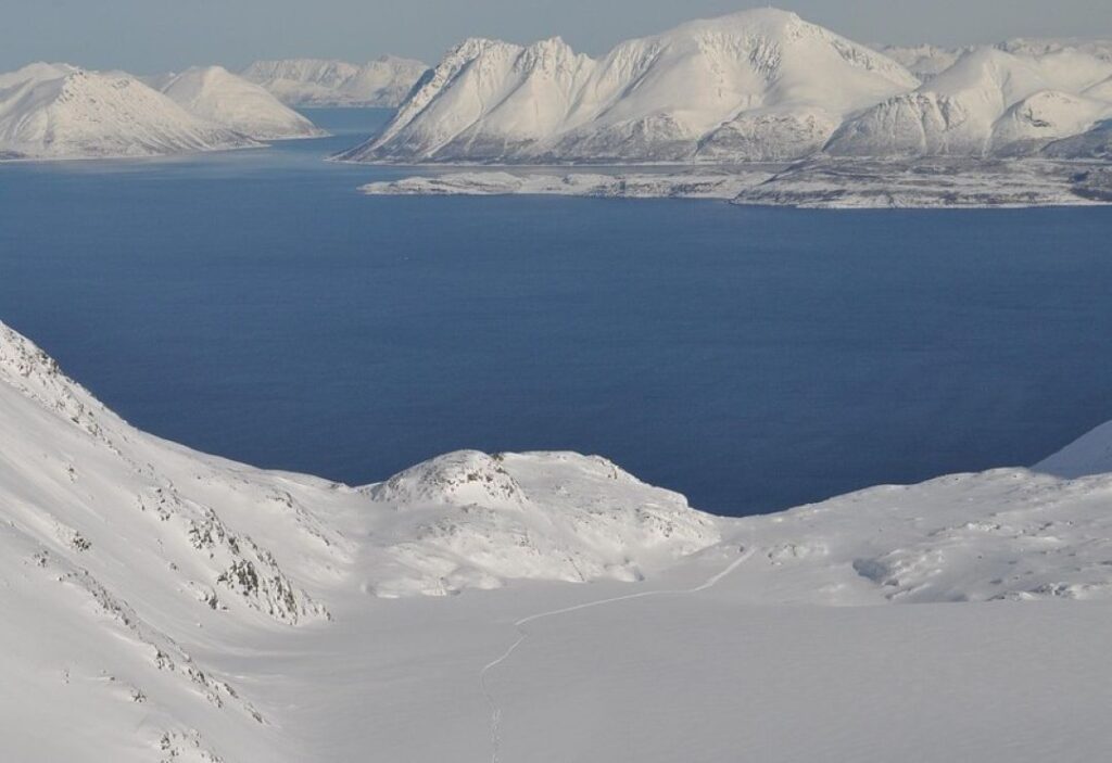 From the top of a snow covered mountain in Arctic norway on a cold bright sunny winter day, with the dark fjord far below, and mountains after mountains stretching indefinitely, covered by white snow glimmering in the sunlight across the fjord