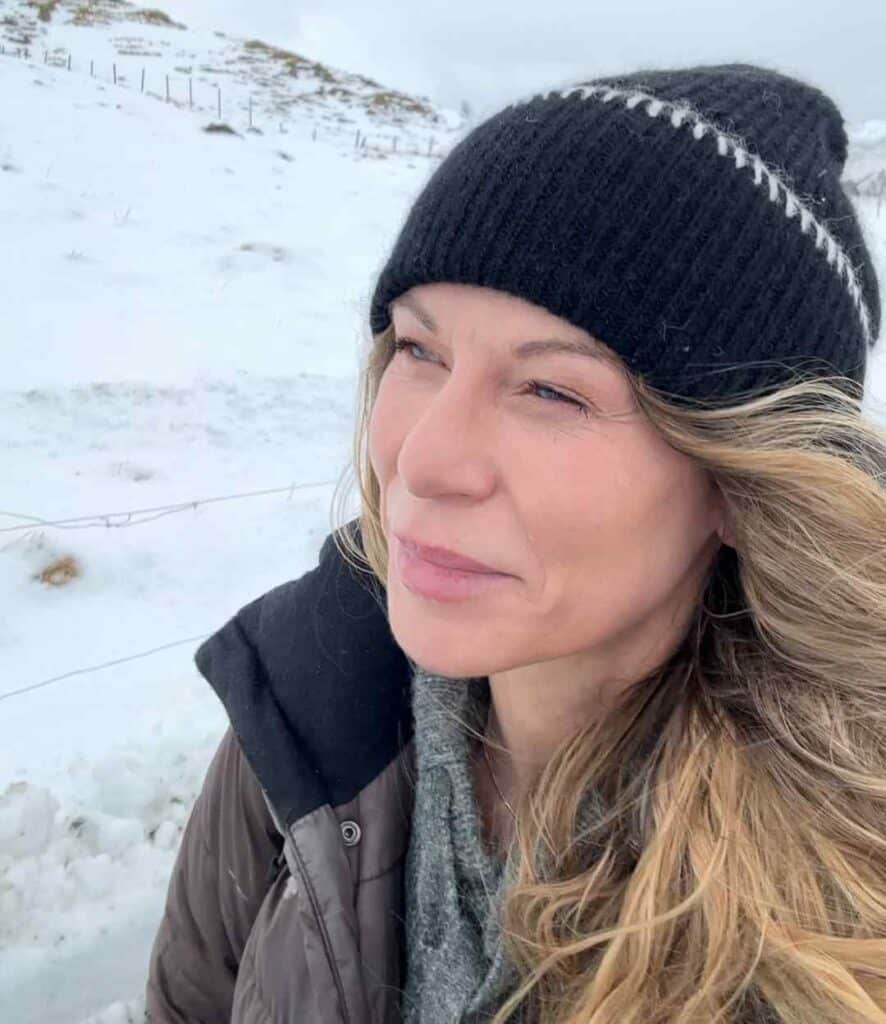 Woman with long blond hair with a black winter hat and thick winter clothes surrounded by nature covered in white snow