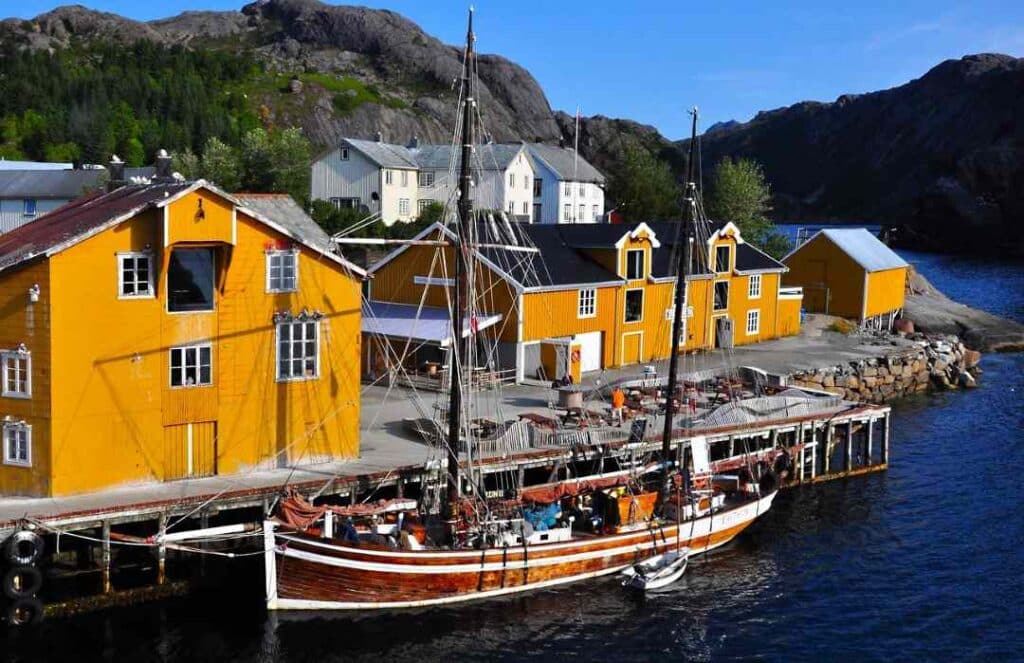 Yellow wooden buildings on the harbor of Nusfjord, a wooden sailboat nestled by the dock on the dark water, and mountains in the background on a sunny summer day
