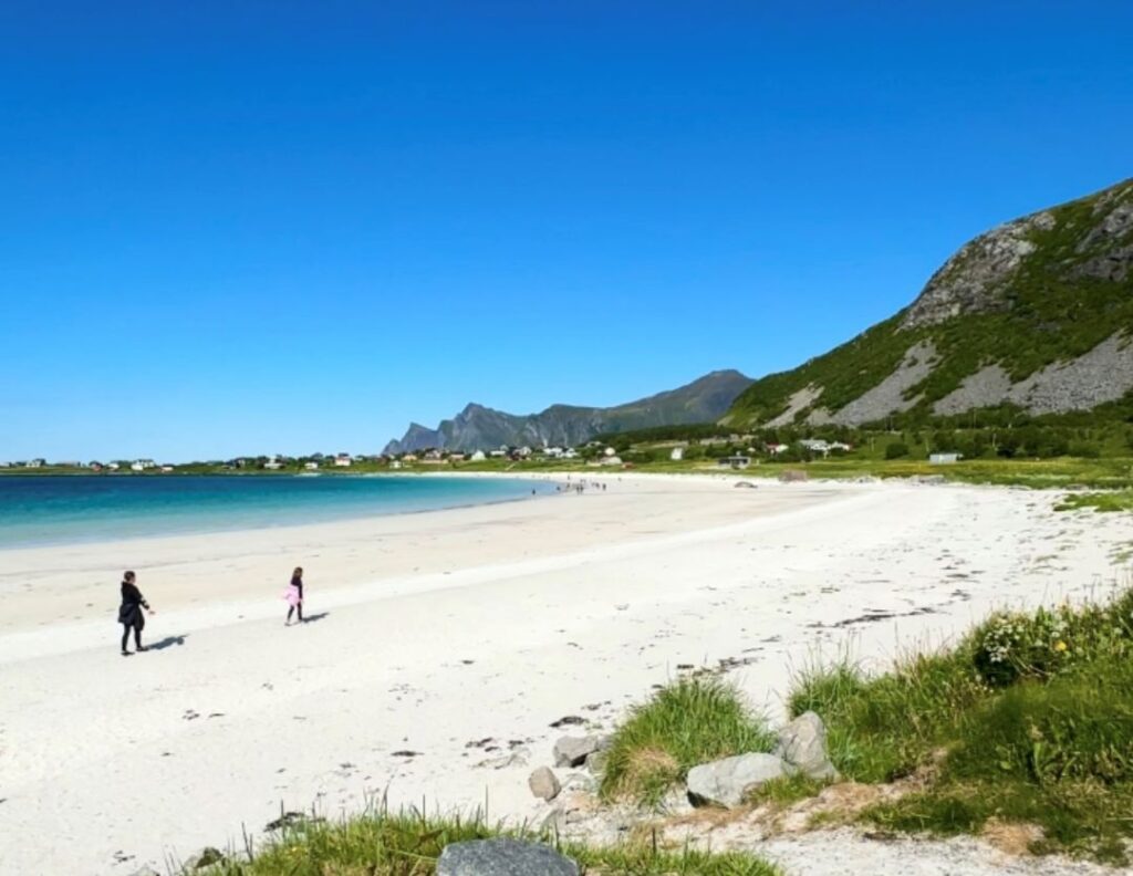 White sandy beach under a clear blue sky, surrounded by green fields and mountains on a sunny day in Ramberg, Lofoten, Norway