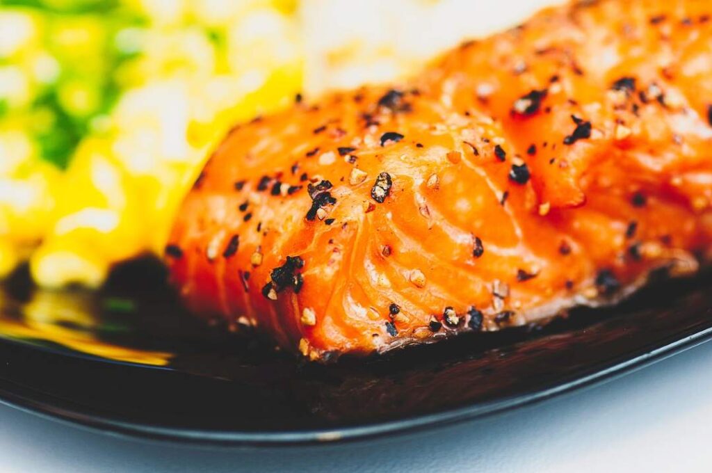 A close up photo of a delicious looking salmon on a black plate, covered in spices, with yellow garniture in the background