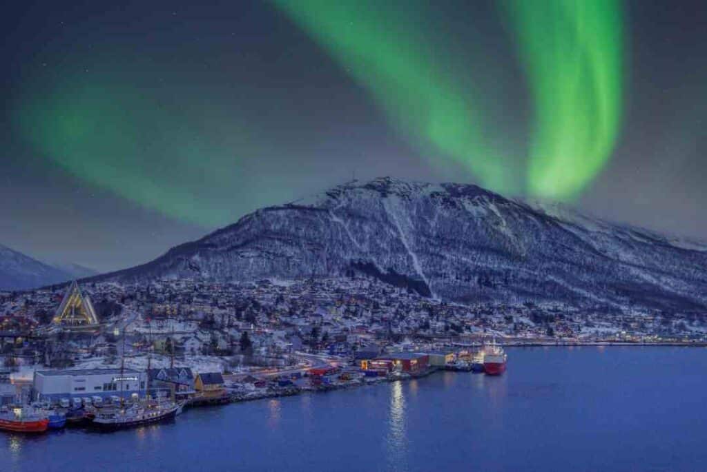 The green aurora borealis dancing on the night sky over a snow capped mountain and dark fjord in Tromsø Norway during winter in the dark blue light that is the Polar Night