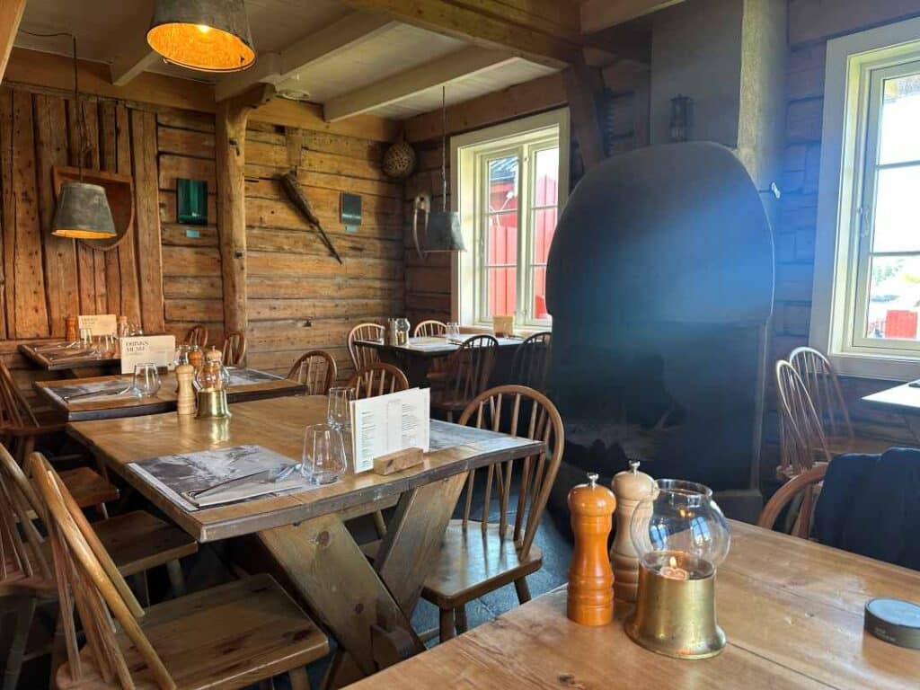 A charming interior in a traditional restaurant, with old log walls, a huge fireplace, and wooden tables and chairs under unique lamps that give a warm light at Gammelbua Restaurant in Reine, Lofoten, Norway. 