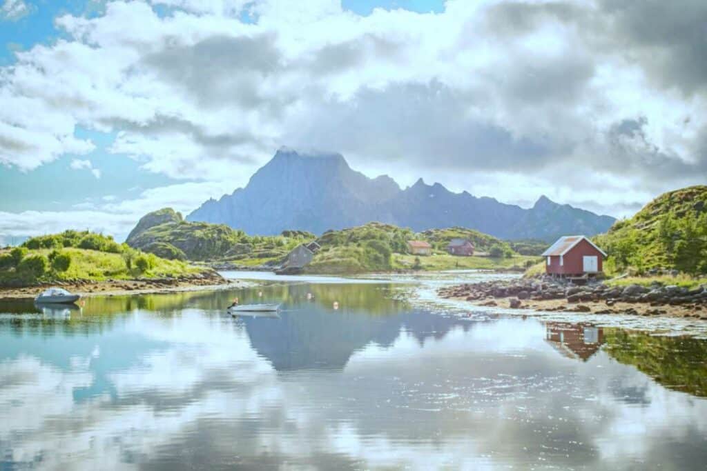A quiet tranquil fjord with a boat on the water and a red cabin in the foreground of majestic mountains on a sunny summer day with green hills, and the sky mirroring in the calm waters in Kabelvåg, Lofoten, Norway