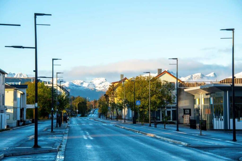 An empty small town main street in the winter, with snow covered mountains in the backgrund, the trees still have some green, under a pale blue winter sky in Leknes, Lofoten, Norway