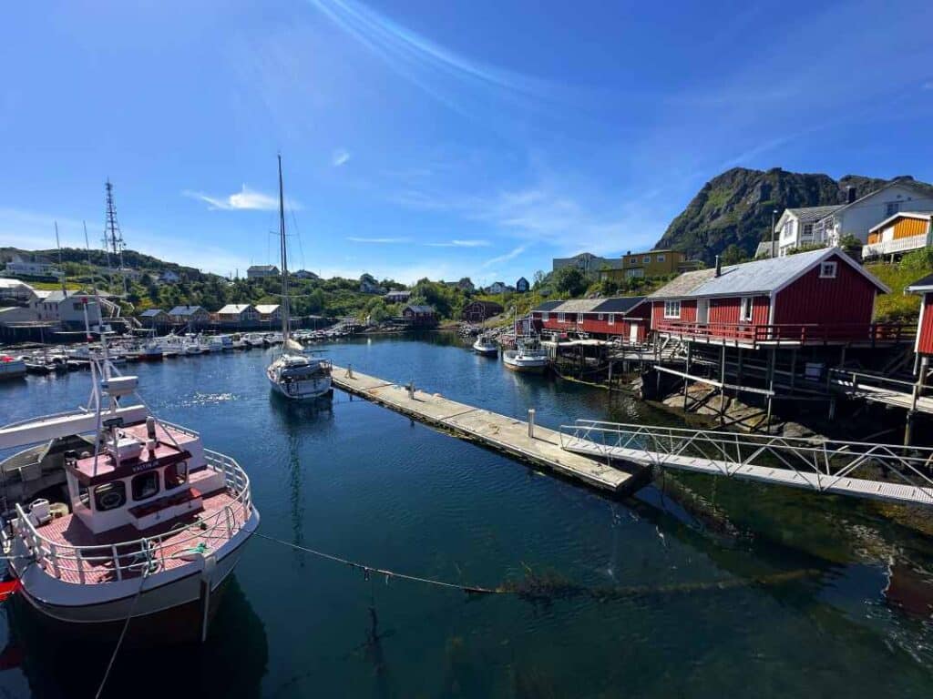 A calm harbor on a sunny summer day with fishing boats scattered around the harbor below red Fishermans cabins under a blue sky, mountains in the distance