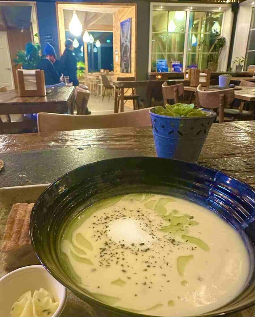 A delicious white fish soup looking elegaint with green spice oils and garniture served in a blue traditional bowl on a rustic wooden table at Anitas Seafood restaurant in Lofoten Norway. The interior around has warm lights, palnts, and charming details. 