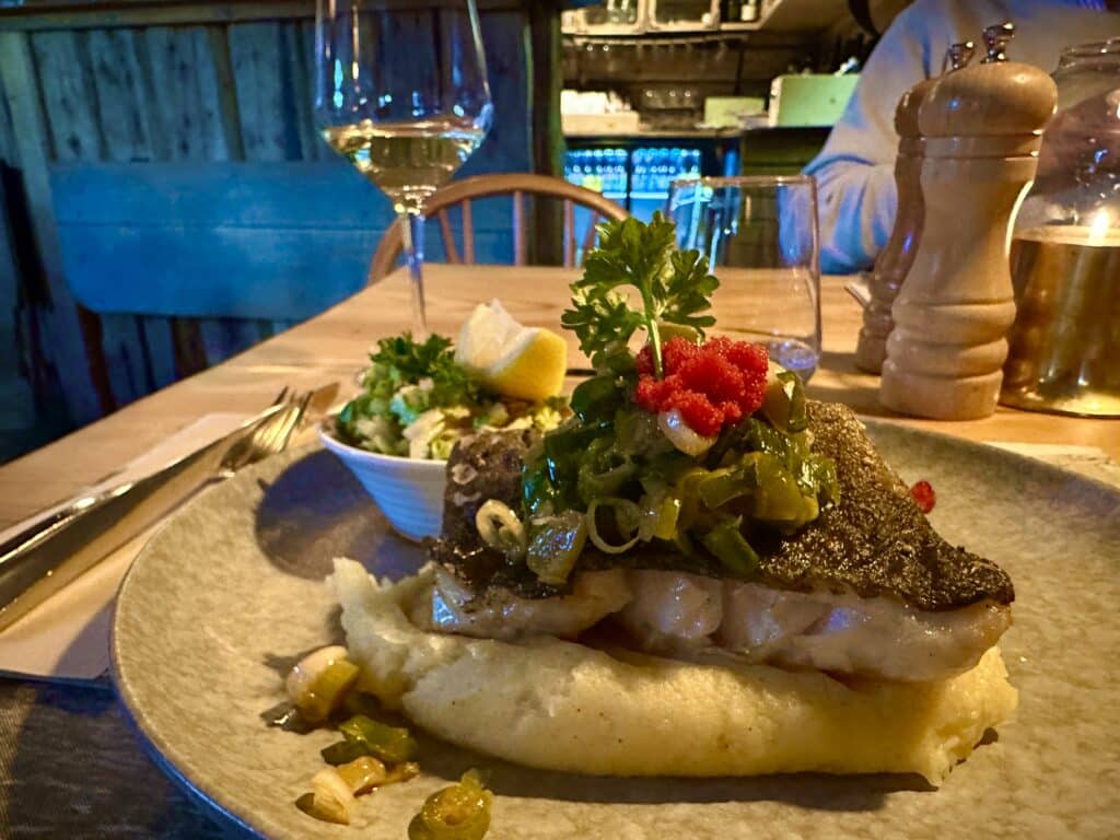 A beautifully presented plate of white fish with fresh vegetables as garniture, a glass of white wine in the background, all in a warm light from an evening lit restaurant in Lofoten, Norway