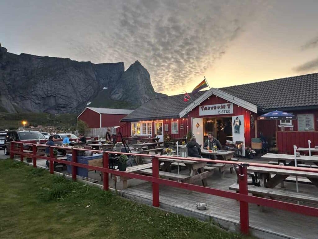 A red traditional wooden house in Reine, Lofoten, which is a restaurant called Tapperiet. Outdoor seating area in front, under a hazy light blue sky at sunset, under majestic mountains of Northern Norway. 