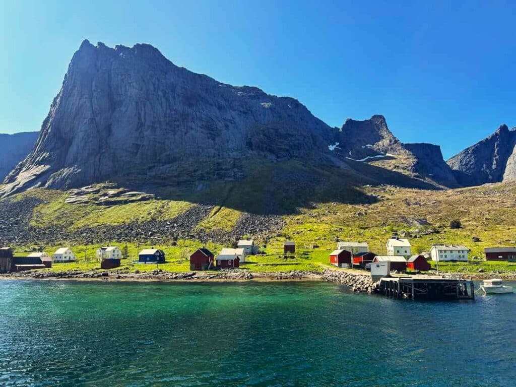 A small settlement of colorful wooden houses in a green pasture under a majestic mountain in Lofoten Norway on a bright sunny day, clear blue skies and the dark greenish blue fjord waters in the foreground