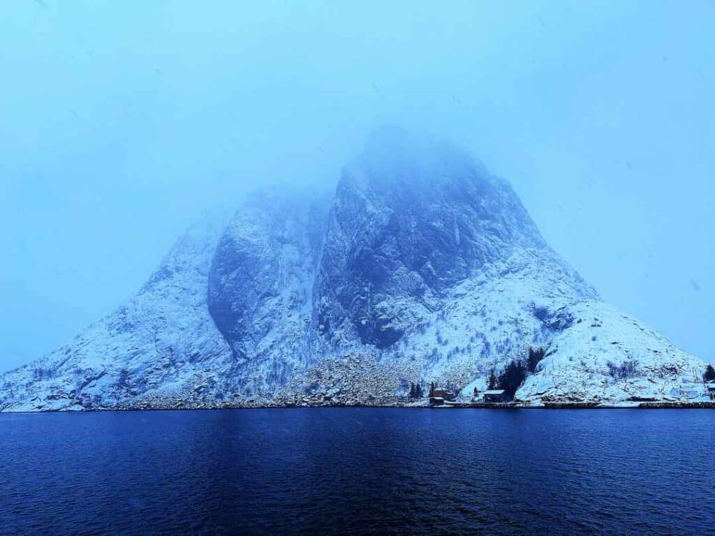 A mountain towering behind a dark fjord, covered in snow and engulfed in light misty clouds at the peak, surrounded by the blue Arctic light in Lofoten Norway
