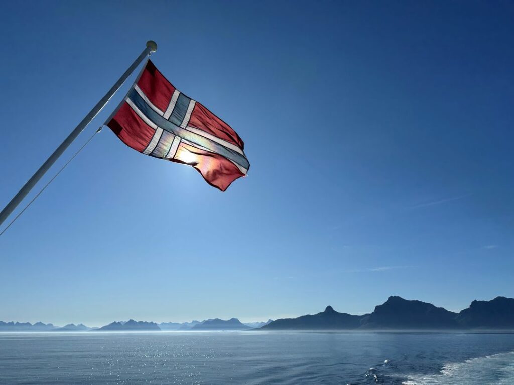 The red white and blue Norwegian flag in the wind on the stern of a ferry from Bodø to Lofoten in Northern Norway on a calm, clear, sunny summer day with silver ocean and blue skies, and the dramatic mountains grey in the distance
