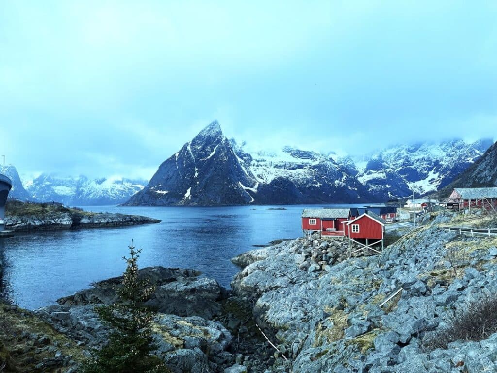 A red rorbu, fishermans cabin, by the fjord surrounded by majestic snow capped mountains under a gloomy autumn sky in Lofoten, Norway